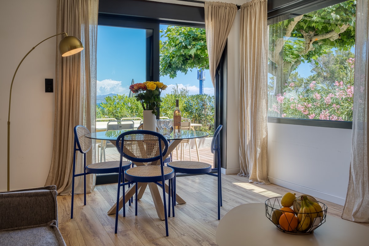A dining area features a glass table surrounded by three elegant chairs, illuminated by natural light. Floor-to-ceiling windows offer views of a verdant exterior and distant ocean, while sheer curtains add a touch of softness to the space. A bowl of fruits rests on a side table.
