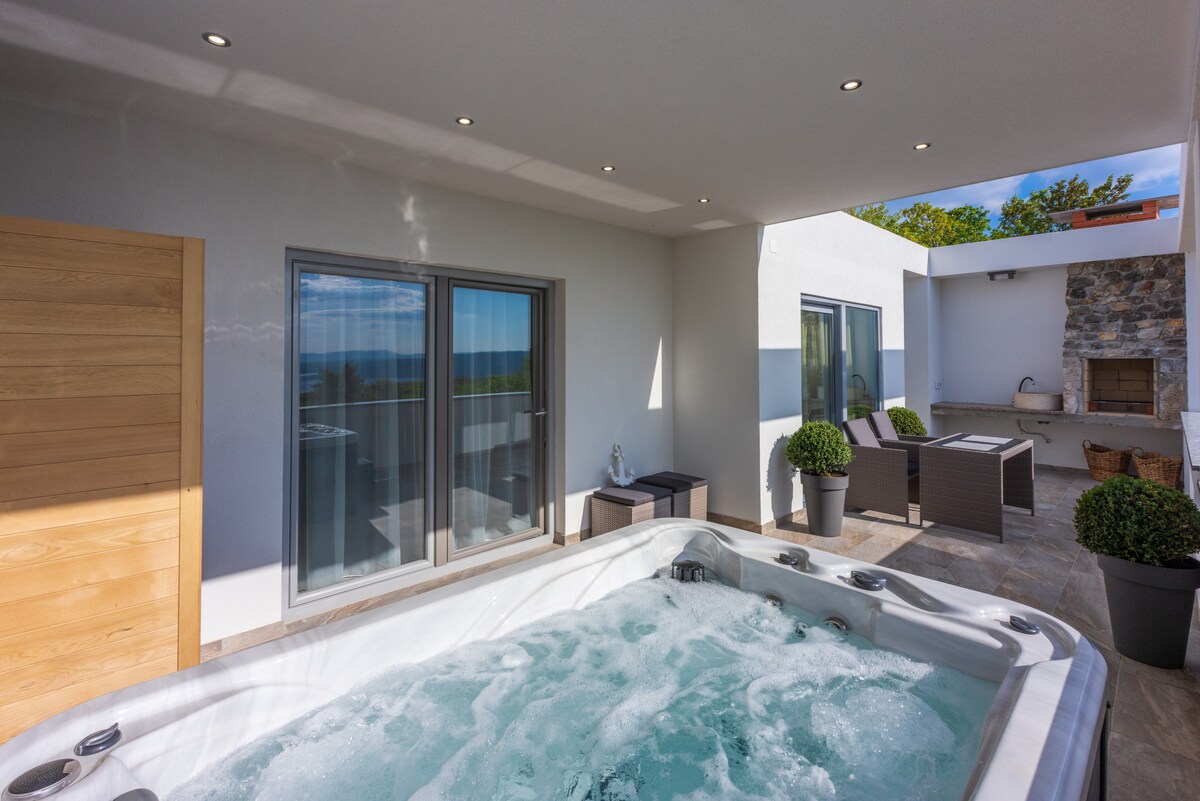 An outdoor terrace features a spacious jacuzzi surrounded by a decorative stone wall. Two large windows offer natural light and views of the surroundings. Comfortable seating arrangements are visible alongside the jacuzzi, enhancing the area for relaxation.