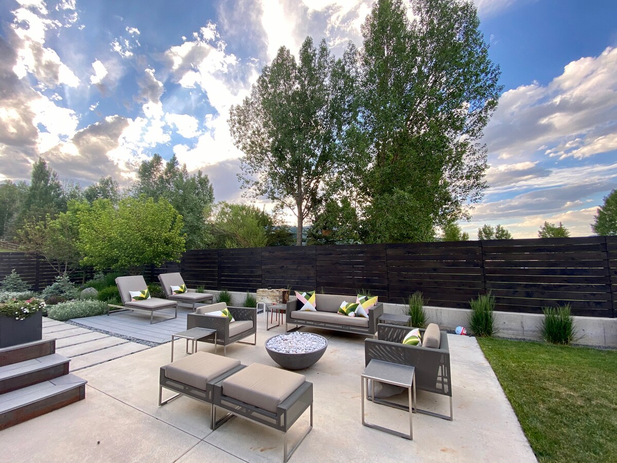 An outdoor seating area features multiple lounge chairs and seating ottomans arranged around a circular fire pit. Lush greenery surrounds the space, with a wooden fence in the background. The sky above showcases scattered clouds, creating a serene atmosphere.