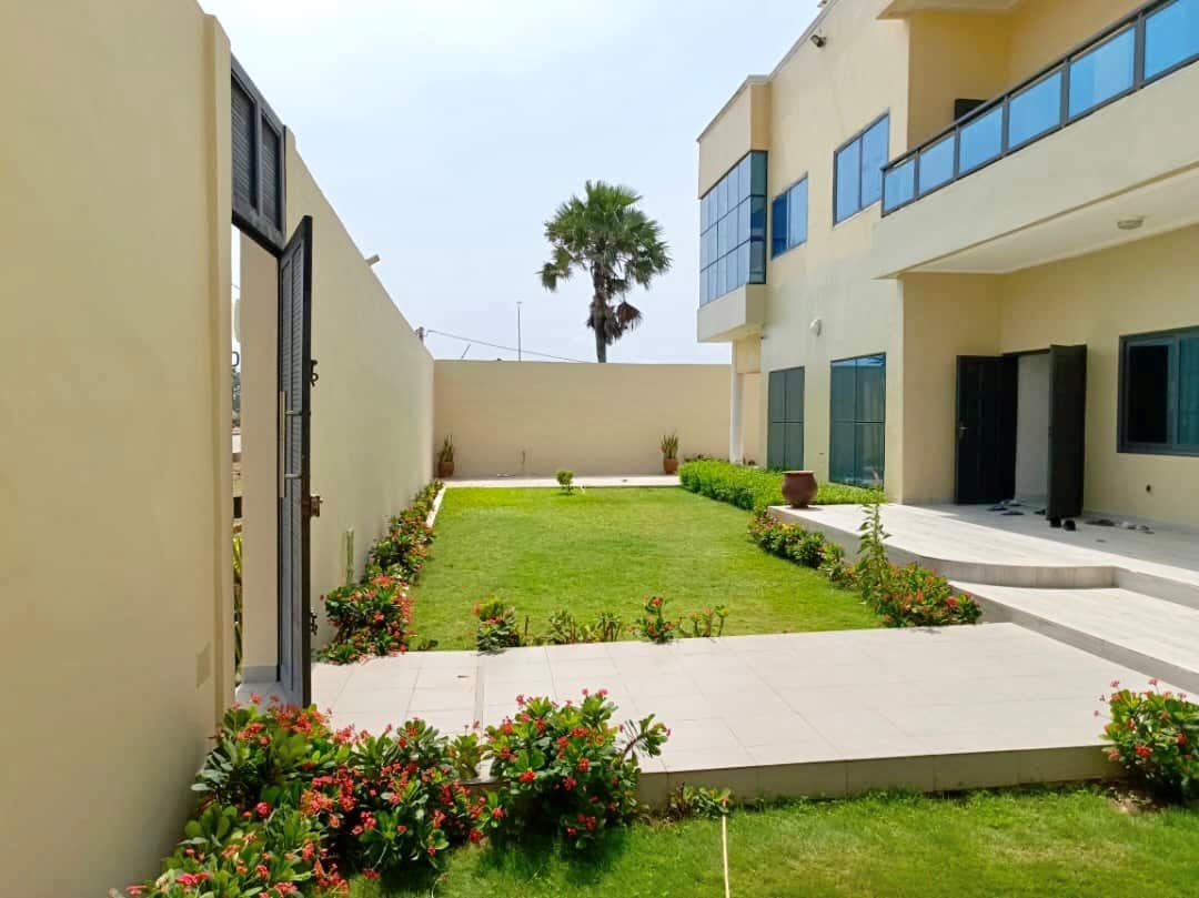 A well-maintained garden is showcased with lush green grass and colorful flower beds lining the pathway. The villa's exterior is visible with large windows and a modern design. A palm tree adds a tropical touch to the serene outdoor space.