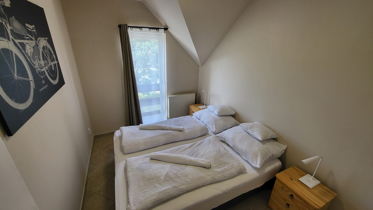 A well-lit sleeping area presents two spacious beds with crisp white bedding. Each bed is complemented by a wooden nightstand, featuring modern reading lamps. A window draped with light curtains allows natural light to fill the room, enhancing the serene ambiance.