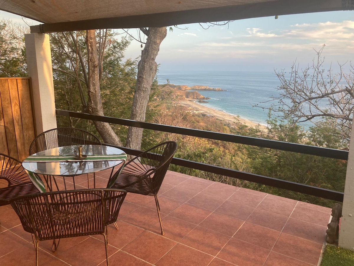 A terrace with a circular glass table and four wicker chairs offers ocean views. The surrounding area is framed by trees, while the coastline is visible in the distance. The warm tones of the tiled floor complement the natural setting.