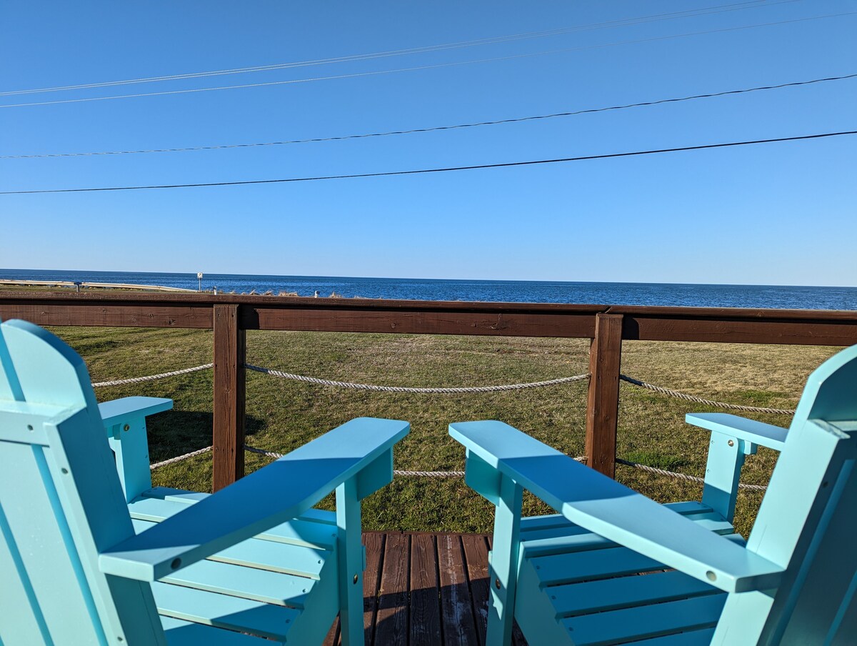 Two light blue Adirondack chairs are positioned on a wooden deck, providing a view of the calm ocean water in the distance. A clear blue sky extends above, with grass visible in the foreground. The scene emphasizes a peaceful outdoor setting.
