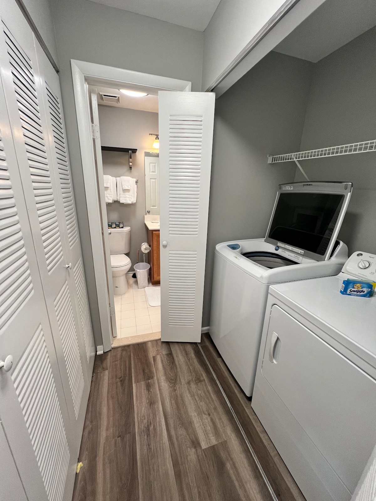 A laundry area features a washing machine and a dryer, with storage space above. To the side, a door leads to a full bathroom, which is equipped with towels and toiletries. Light wood flooring connects the two spaces, creating a functional layout.
