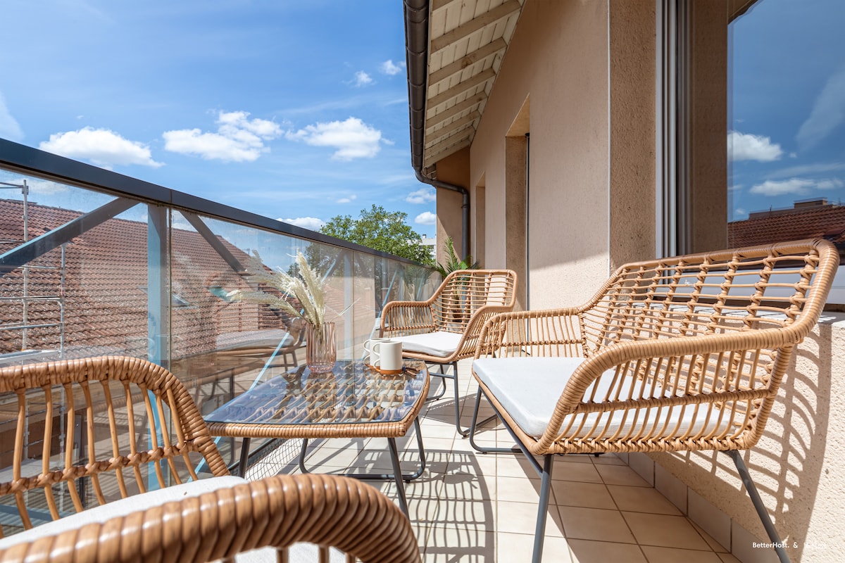A balcony is presented with two wicker chairs and a matching table, inviting relaxation. The space is bathed in natural light, with a clear view of the sky and surrounding rooftops. Greenery is placed on the table, enhancing the serene atmosphere.