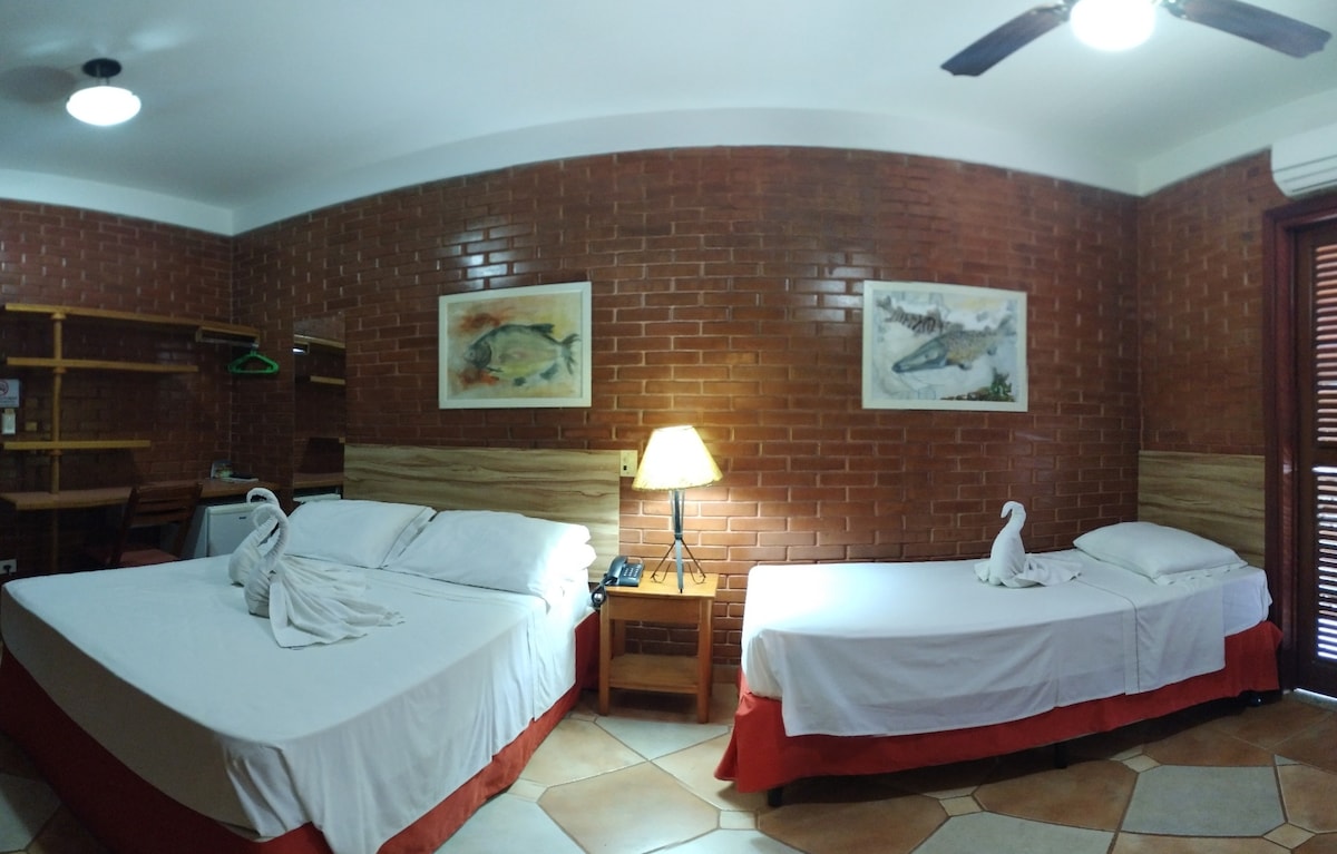 The guest room features two beds arranged with white linens, one being a spacious bed and the other a single. A ceiling fan and bedside lamp provide lighting. Artwork hangs on exposed brick walls, and wooden shelving offers storage options.