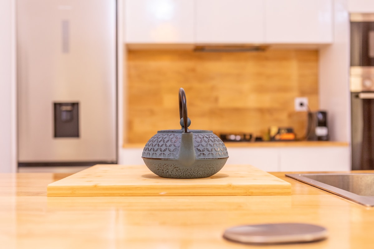 A textured kettle is positioned on a wooden cutting board in the modern kitchen. The kitchen features sleek cabinetry and stainless steel appliances in the background, contributing to an organized cooking space.