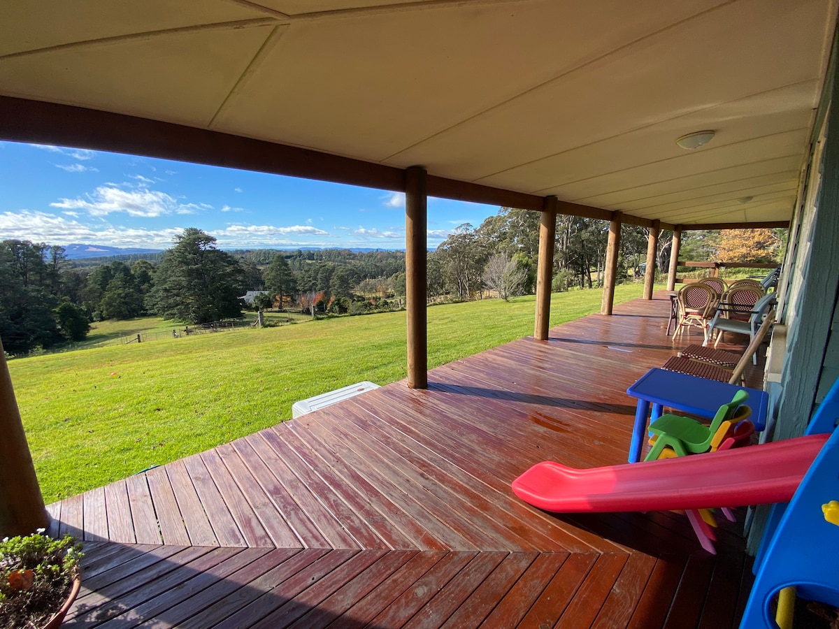 An expansive back verandah features polished wooden flooring and sturdy posts. Colorful play equipment is visible on one side, alongside a set of outdoor chairs and a small table, overlooking a lush green vista of rolling hills and distant mountains under a clear blue sky.