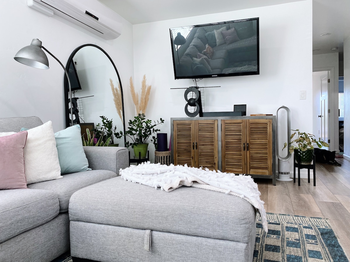 A cozy living room setting features a large gray sectional couch adorned with pastel cushions. A round mirror and a modern floor lamp enhance the space, while a wooden cabinet rests against the wall, complemented by a flat-screen TV. Decorative plants provide a touch of greenery.