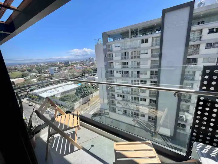A balcony view presents two wooden chairs positioned on a sleek surface. The scene offers a glimpse of the surrounding cityscape and nearby buildings, with expansive skies above and hints of distant mountains in the background.