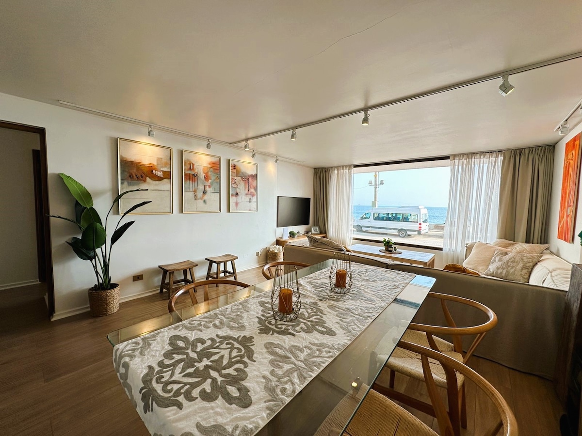 The living area features a large window providing views of the sea, with natural light illuminating the room. A comfortable sofa and stylish decor are present, along with three wooden stools and a dining table set with a decorative table runner. Attractive artwork adorns the walls.