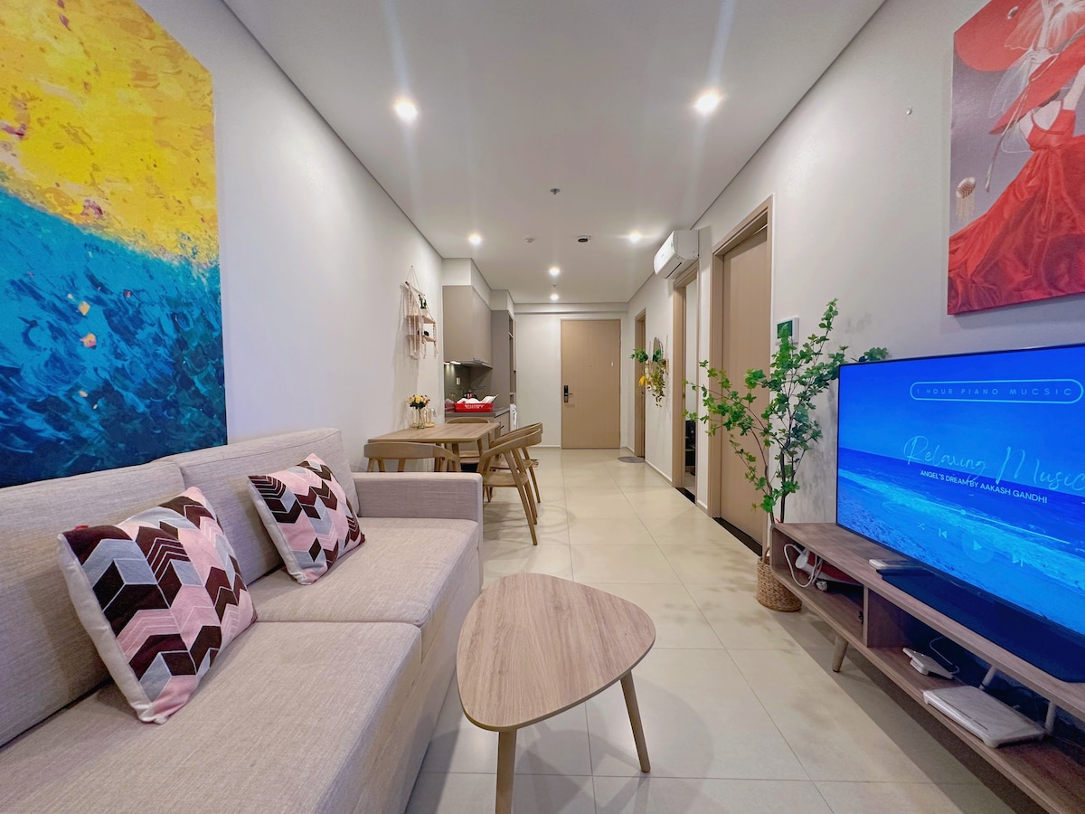 A spacious living area features a light-colored sofa adorned with patterned cushions. A wooden coffee table sits in front, and a dining table with chairs is visible in the background. A television is positioned on a console against the wall, complemented by wall art and greenery.