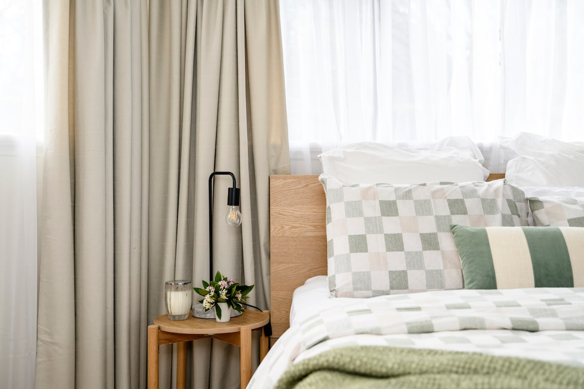 A spacious bed is paired with a patterned blanket and an assortment of decorative pillows. A wooden side table holds a vase with fresh flowers and a softly glowing light fixture. Natural light is filtered through sheer curtains, creating a calm atmosphere.