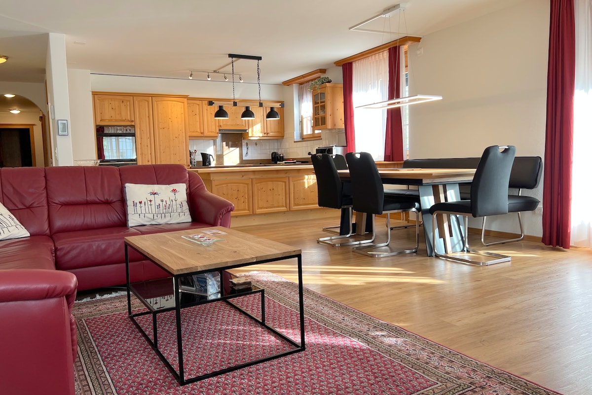 The spacious living area includes a large red sofa facing a wooden coffee table on a patterned rug. A dining table with black chairs is present, illuminated by natural light from large windows. An open kitchen with wooden cabinetry is visible in the background.