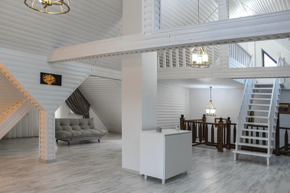 The open-concept interior features a spacious layout with white wooden paneling and vaulted ceilings. A cozy seating area with a light-colored sofa is visible, alongside a sleek white cabinet. Light fixtures hang from the ceiling, enhancing the airy atmosphere.