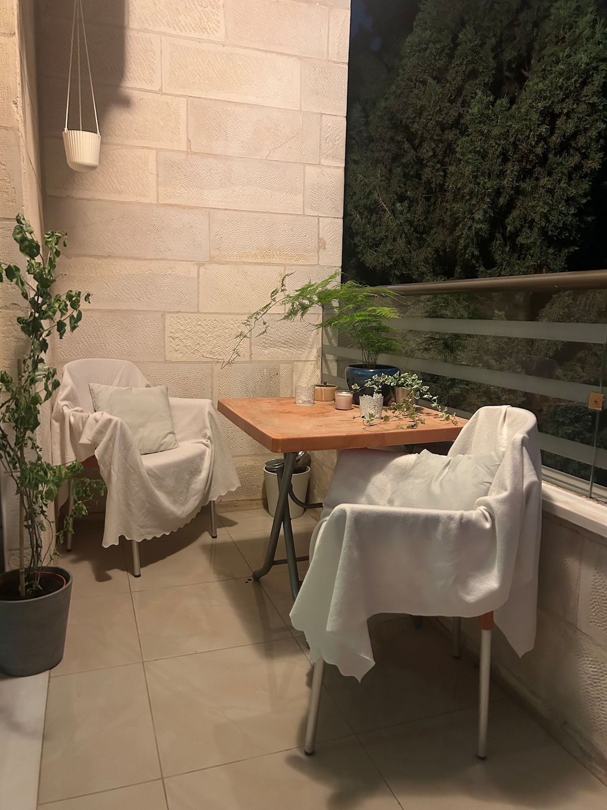 A cozy balcony is presented with two white chairs and a small wooden table, softly illuminated at night. Green plants add a touch of nature, while a light drape partially covers the chairs. A railing provides a view of the surrounding trees.
