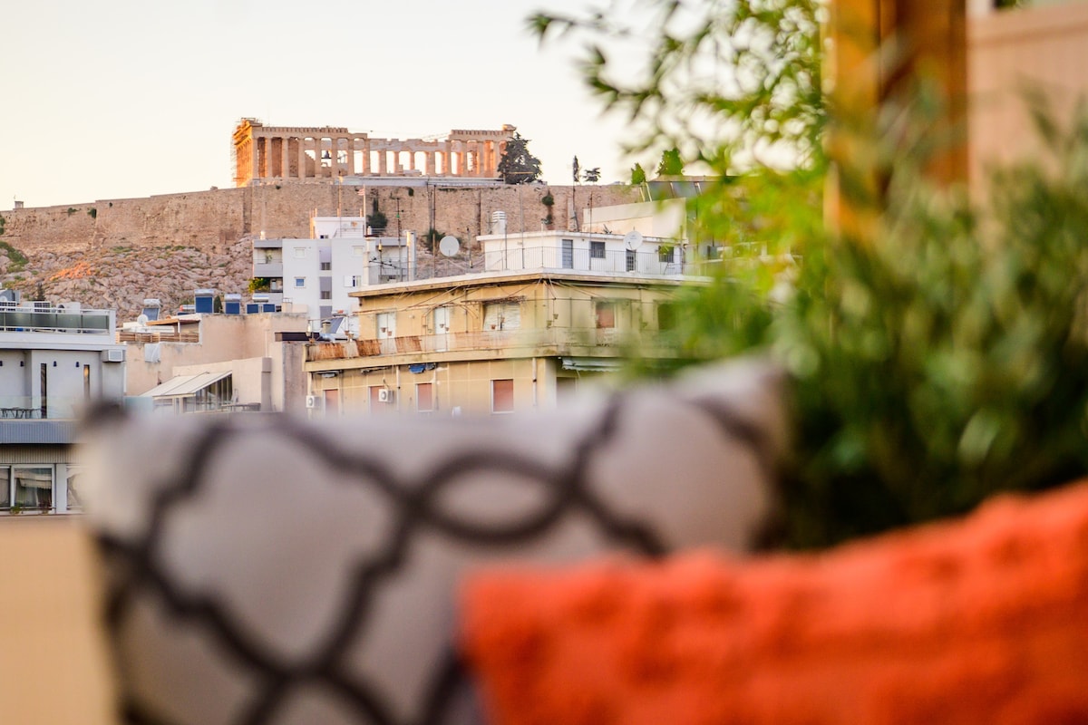 A cozy outdoor daybed is visible in the foreground, adorned with patterned cushions. In the background, the Acropolis is showcased atop a distant hill, illuminated by the soft glow of sunset, amidst a backdrop of city buildings.