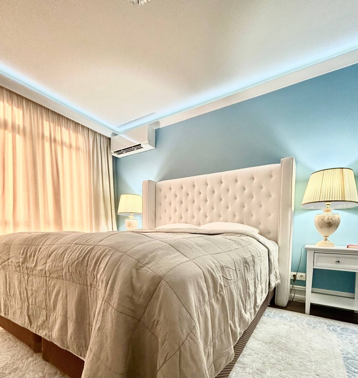 A well-appointed bedroom features a spacious bed with an upholstered headboard. Soft light from two bedside lamps adds warmth to the room, while curtains gently filter natural light from a window. A light blue wall complements the neutral tones of the bedding and carpet.