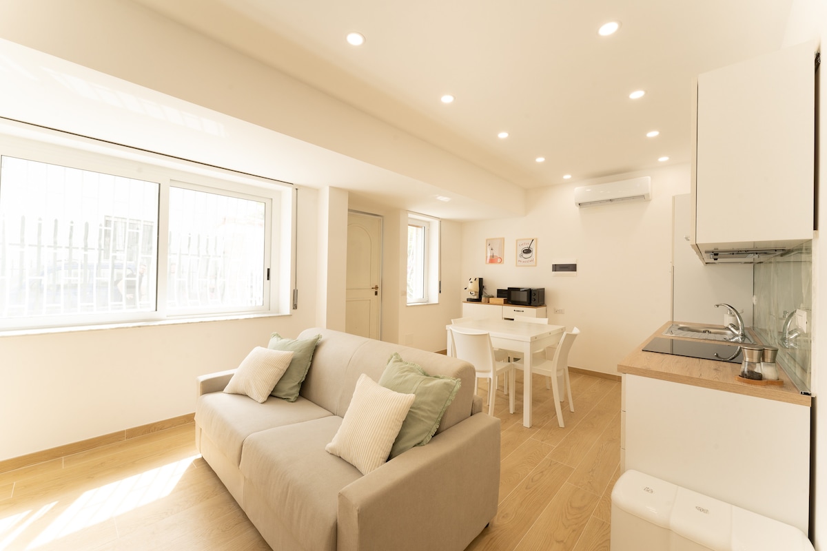 A bright living area is illuminated by ceiling lights and features a cozy sofa with decorative cushions. A dining table with chairs is positioned nearby, along with a modern kitchen equipped with essential appliances. Large windows allow natural light to fill the space.