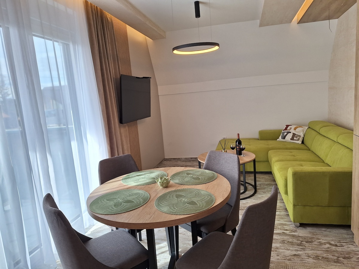 A modern living area showcases a green sectional sofa next to a round dining table set with placemats. Two gray chairs surround the table, which is complemented by a small decorative bowl. Large windows allow natural light to illuminate the space.