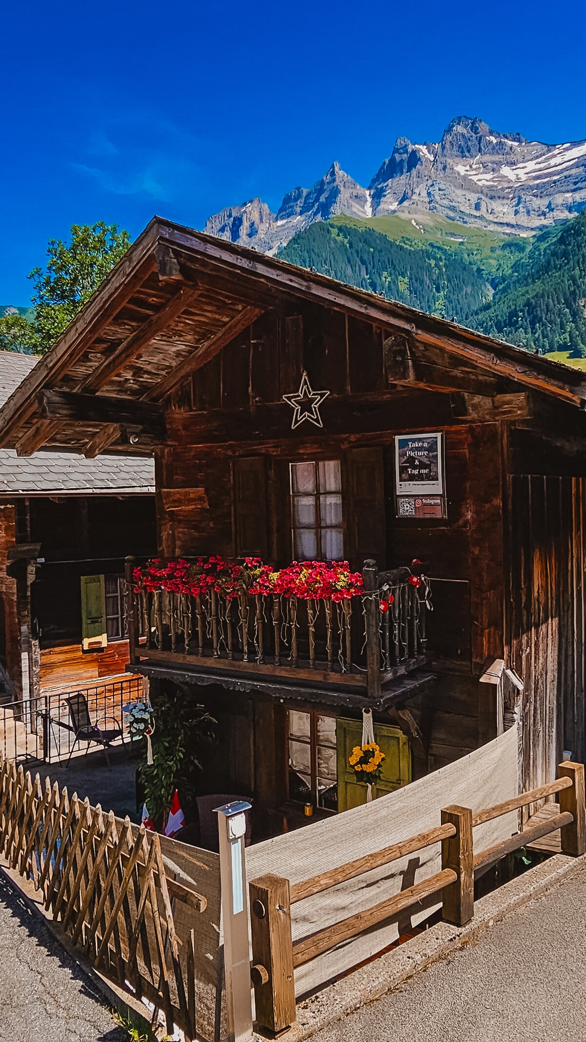 A rustic alpine house features a wooden facade and decorative flower boxes filled with vibrant blooms. A small balcony overlooks the surroundings, framed by a wooden railing. Majestic mountains rise in the background under a clear blue sky, enhancing the serene mountain atmosphere.