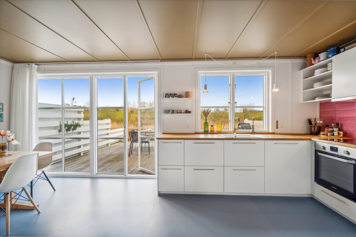 A modern kitchen is shown, featuring sleek white cabinetry and a wooden countertop. Large glass doors open to a deck outside, allowing natural light to fill the space. A dining area with chairs is visible, complemented by soft natural tones and decorative items on the shelves.