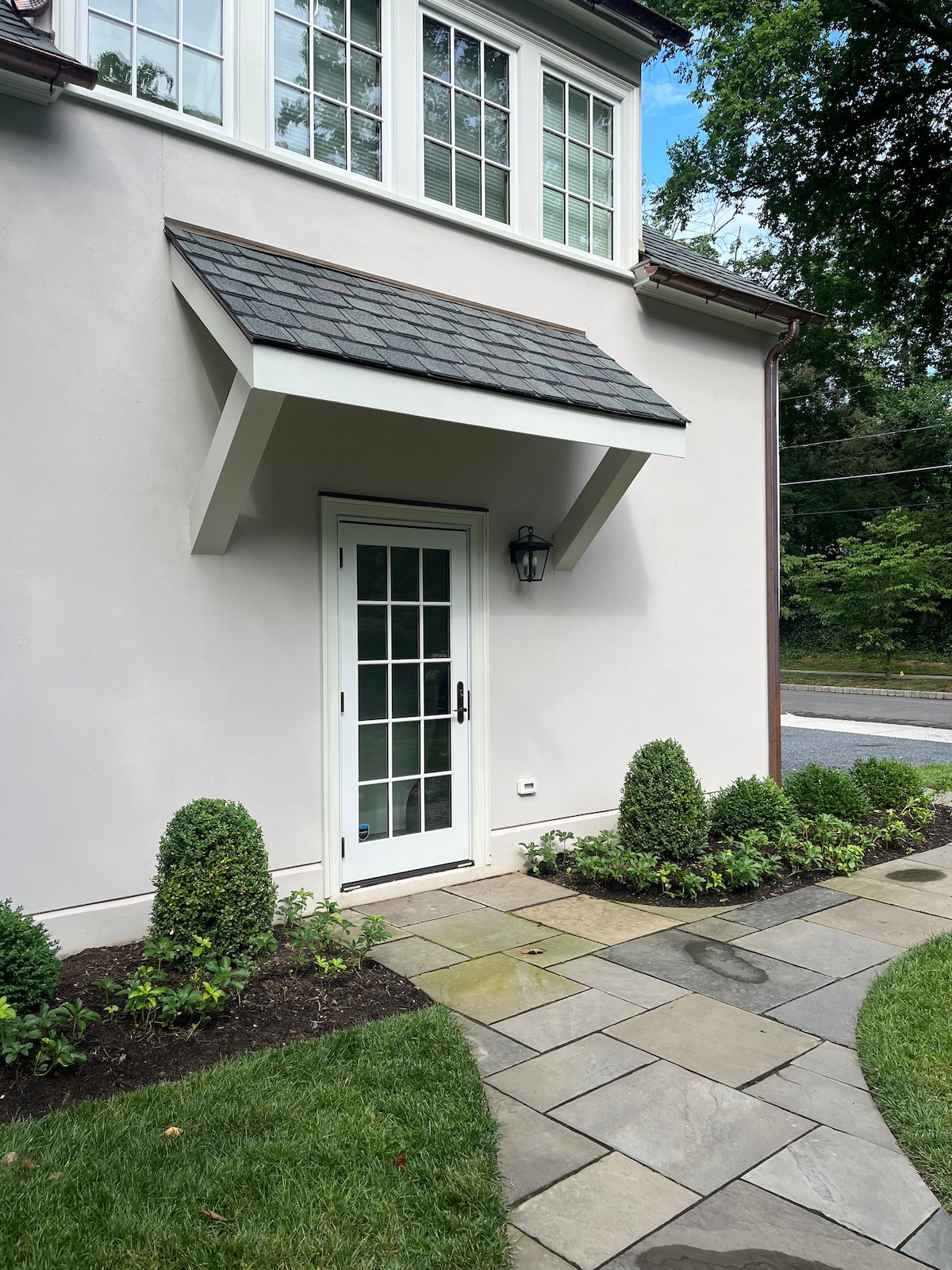 The entrance to the studio apartment features a white door with a glass panel, framed by a decorative awning. Neatly trimmed shrubs are planted along the stone pathway, which leads to the door. The approach is lined with manicured grass, adding to the inviting entry.