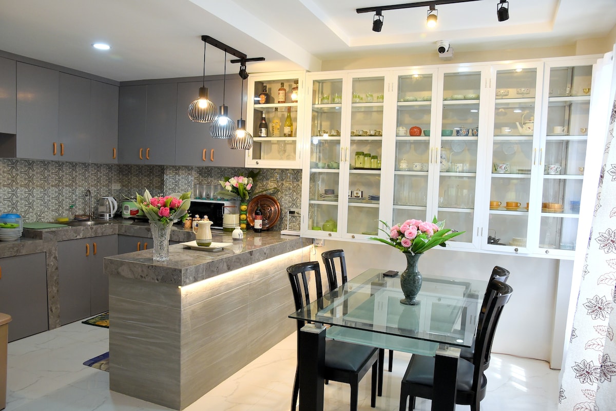 A modern kitchen and dining area is displayed, featuring stylish cabinetry and a glass-top dining table surrounded by black chairs. Natural light illuminates the space, highlighting decorative glassware in the upper cabinets. Fresh flowers are arranged in a vase on the table.