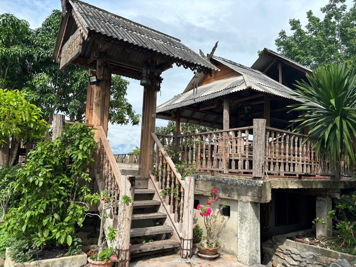 The exterior of the country house is showcased, featuring wooden steps leading to a raised entrance. A wraparound balcony is lined with sturdy railings and adorned with tropical plants, while the roof is designed with traditional thatch, enhancing the natural surroundings.