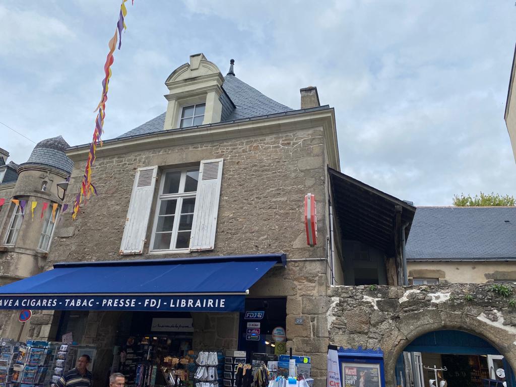 A charming stone building is displayed, showcasing a distinctive peaked roof and large white shutters. Below, a blue awning indicates a shop with signage for tobacco, press, and library services. Colorful flags are seen adorning the exterior, contributing to the vibrant street scene.