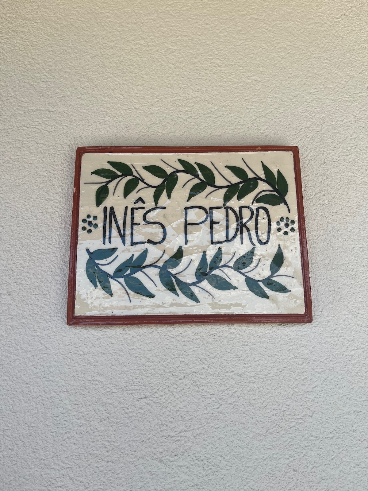 A decorative wall plaque features the names 'Inés' and 'Pedro' surrounded by green leaf motifs. The plaque rests against a light-colored wall, highlighting its intricate design and craftsmanship.