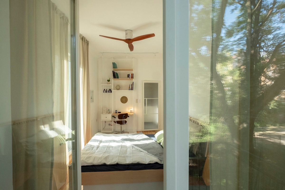 A bright room is visible through sheer curtains, showcasing a double bed with neatly arranged linens. A ceiling fan is installed above, and a work desk with a chair is positioned against the wall. Natural light filters through, illuminating the inviting space.