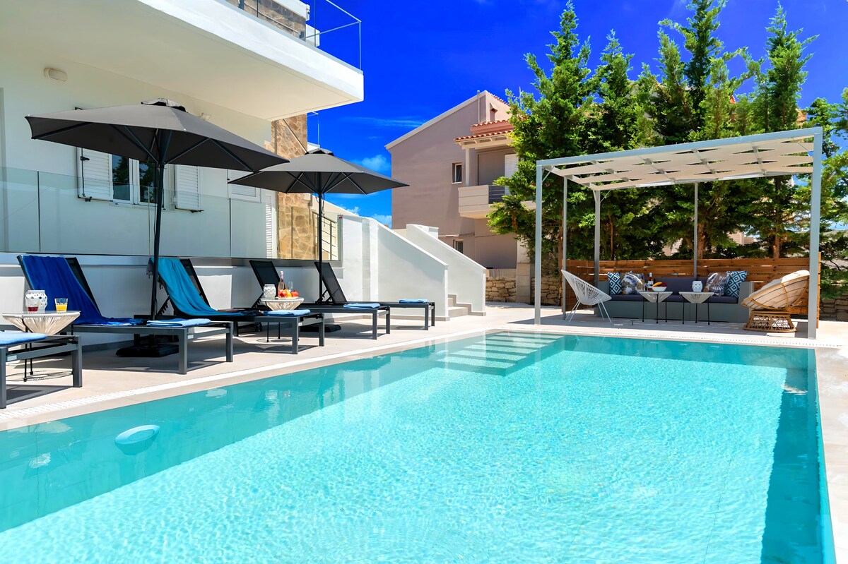 A private swimming pool is surrounded by sun loungers with umbrellas for shade. An outdoor seating area features a comfortable arrangement under a canopy, with greenery providing privacy. The clear water of the pool reflects the bright blue sky, inviting relaxation.