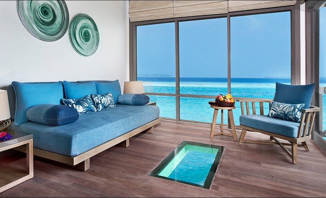A spacious seating area features a large blue sofa adorned with decorative pillows. A wooden chair complements a round table with a fruit arrangement. Floor-to-ceiling windows provide views of turquoise waters, while a glass panel in the floor offers a glimpse into the aquatic environment beneath.