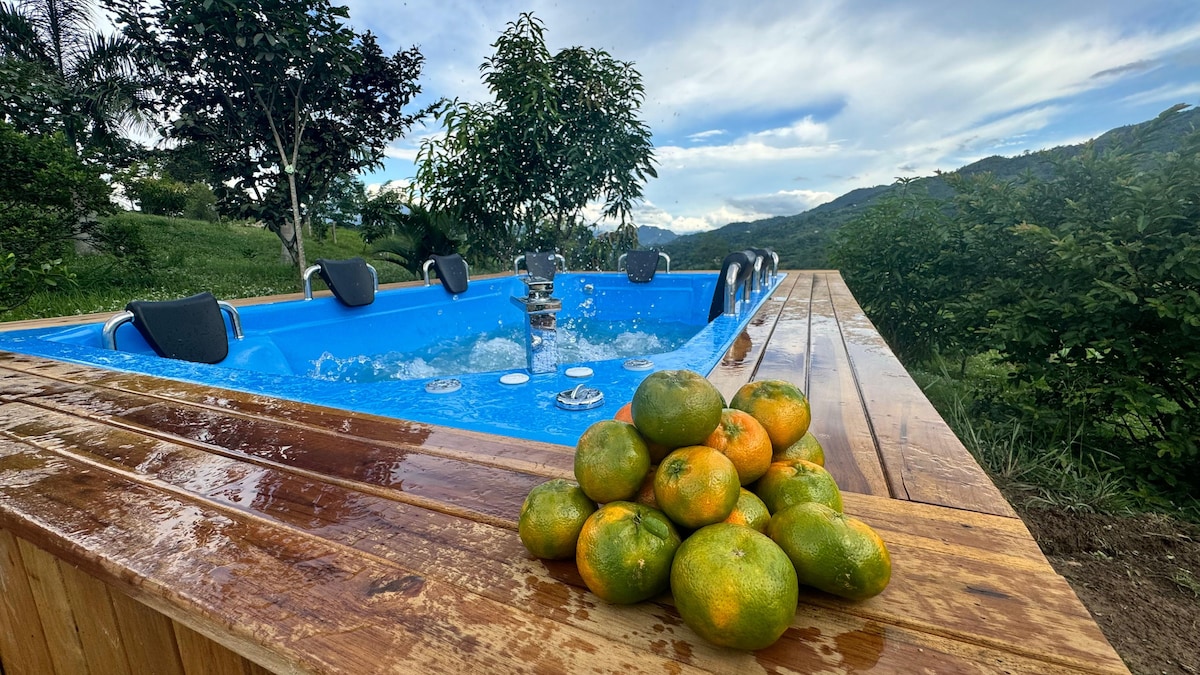 An outdoor jacuzzi is set against a backdrop of rolling hills and greenery, featuring bubbling water and comfortable seating. In the foreground, a pile of fresh oranges rests on a wooden table, highlighting the natural setting of the property.