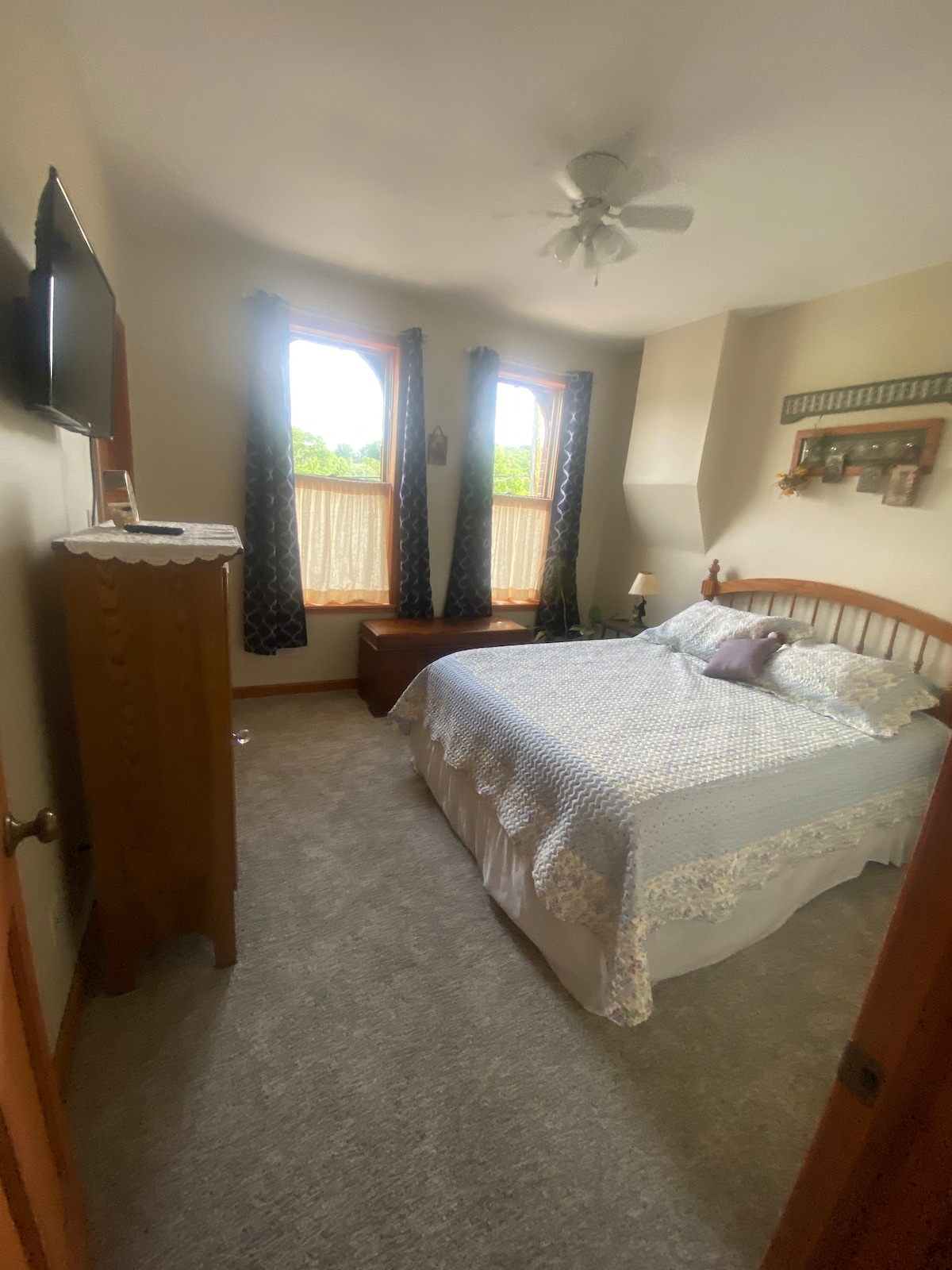 A cozy bedroom is furnished with a queen bed, adorned with a patterned bedspread. Natural light filters through the large windows, framed by dark curtains. A dresser and small bench are visible, enhancing the room’s functionality.