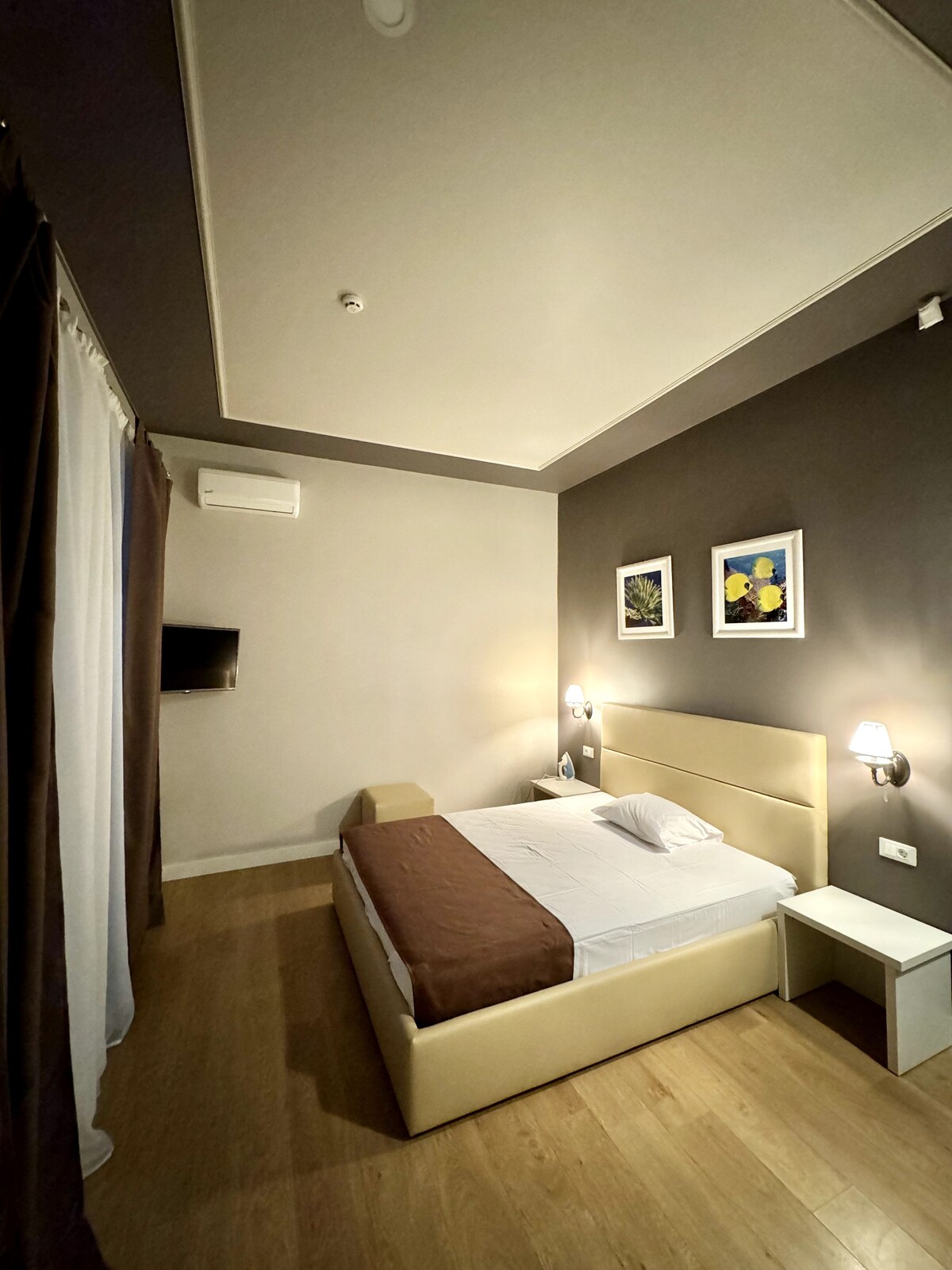 A modern bedroom is showcased with warm grey walls and wooden flooring. A spacious bed is centered against the wall, complemented by nightstands and bedside lamps. Artwork is displayed above the bed, and a wall-mounted TV is visible, providing contemporary amenities.