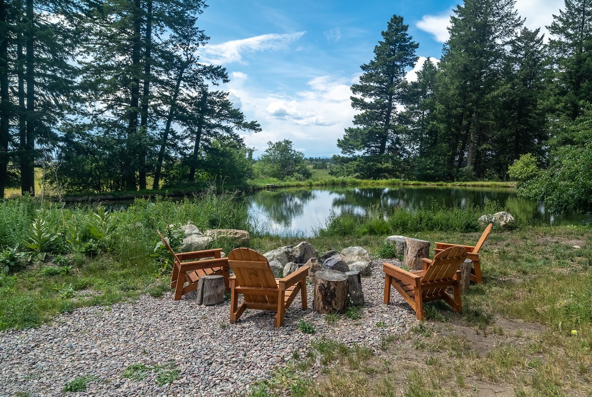 A serene seating area features four wooden chairs arranged around a fire pit made of stones. Nearby, a calm pond reflects the surrounding trees and sky, with grasses and plants enhancing the natural setting.