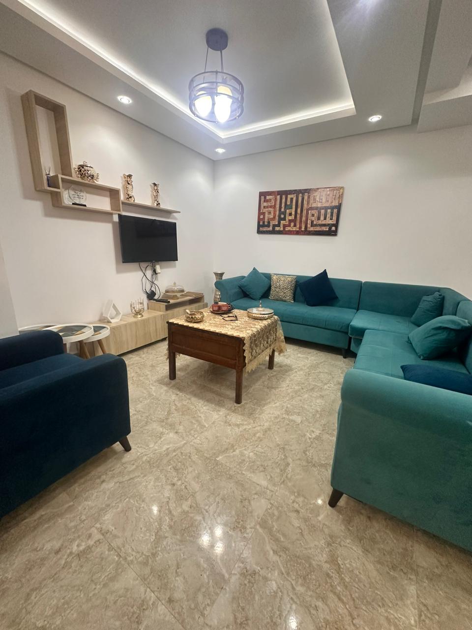 A modern living room features a U-shaped teal sofa complemented by dark blue armchairs. A wooden coffee table with decorative items occupies the center. Art is displayed on the wall, while a flat-screen TV is mounted above a light wooden shelf.