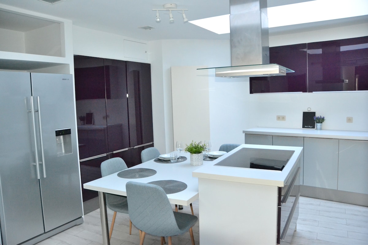 A modern kitchen is equipped with sleek, dark cabinetry and a bright countertop. A central cooking island features a stovetop and is surrounded by a dining table set with four grey chairs. Natural light is welcomed through a skylight above.