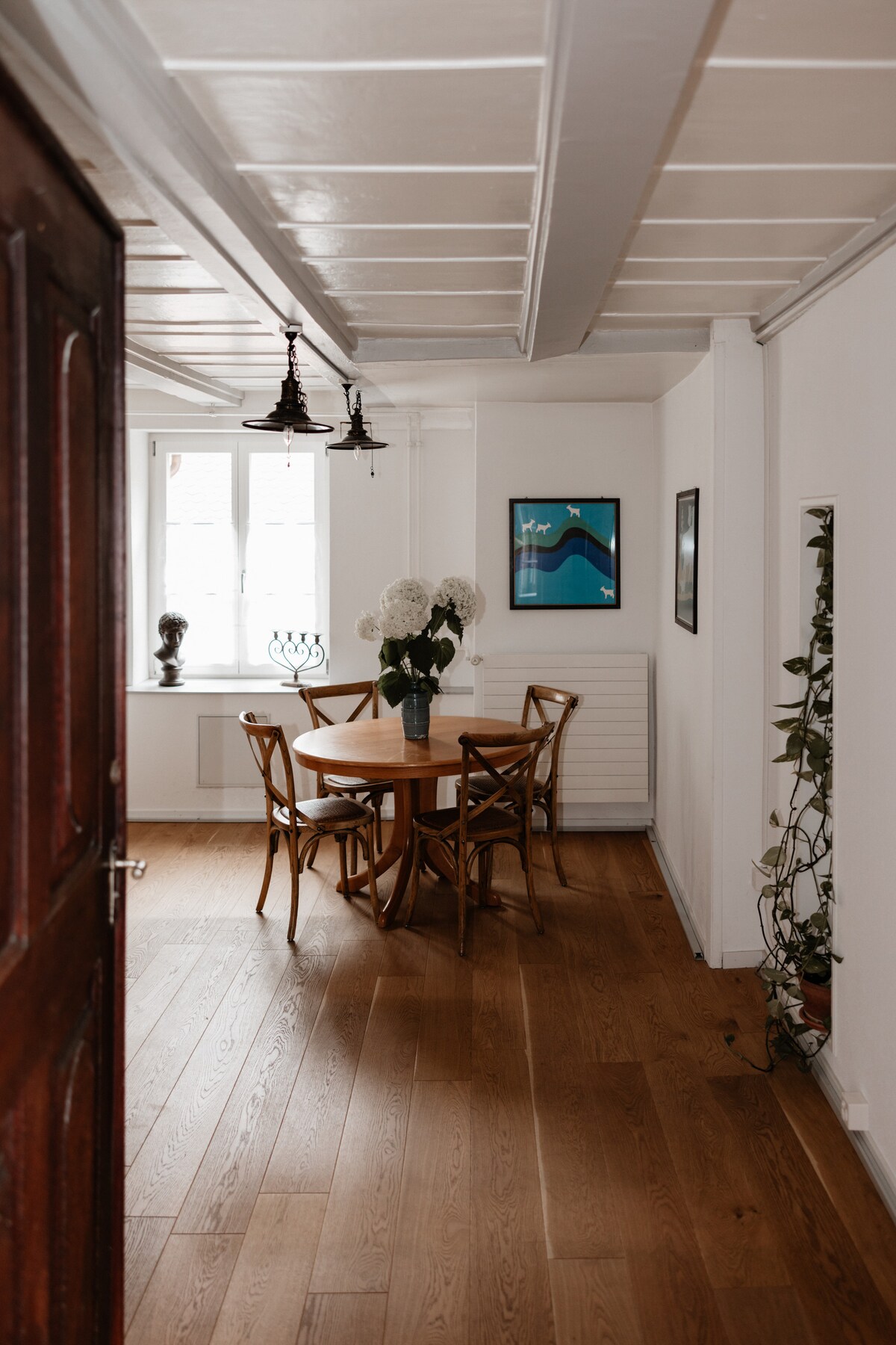A bright dining area features a round wooden table surrounded by four cross-back chairs. Natural light enters through large windows, illuminating the space, while wall-mounted decor adds subtle character. A vase of white flowers sits at the center of the table, enhancing the welcoming atmosphere.