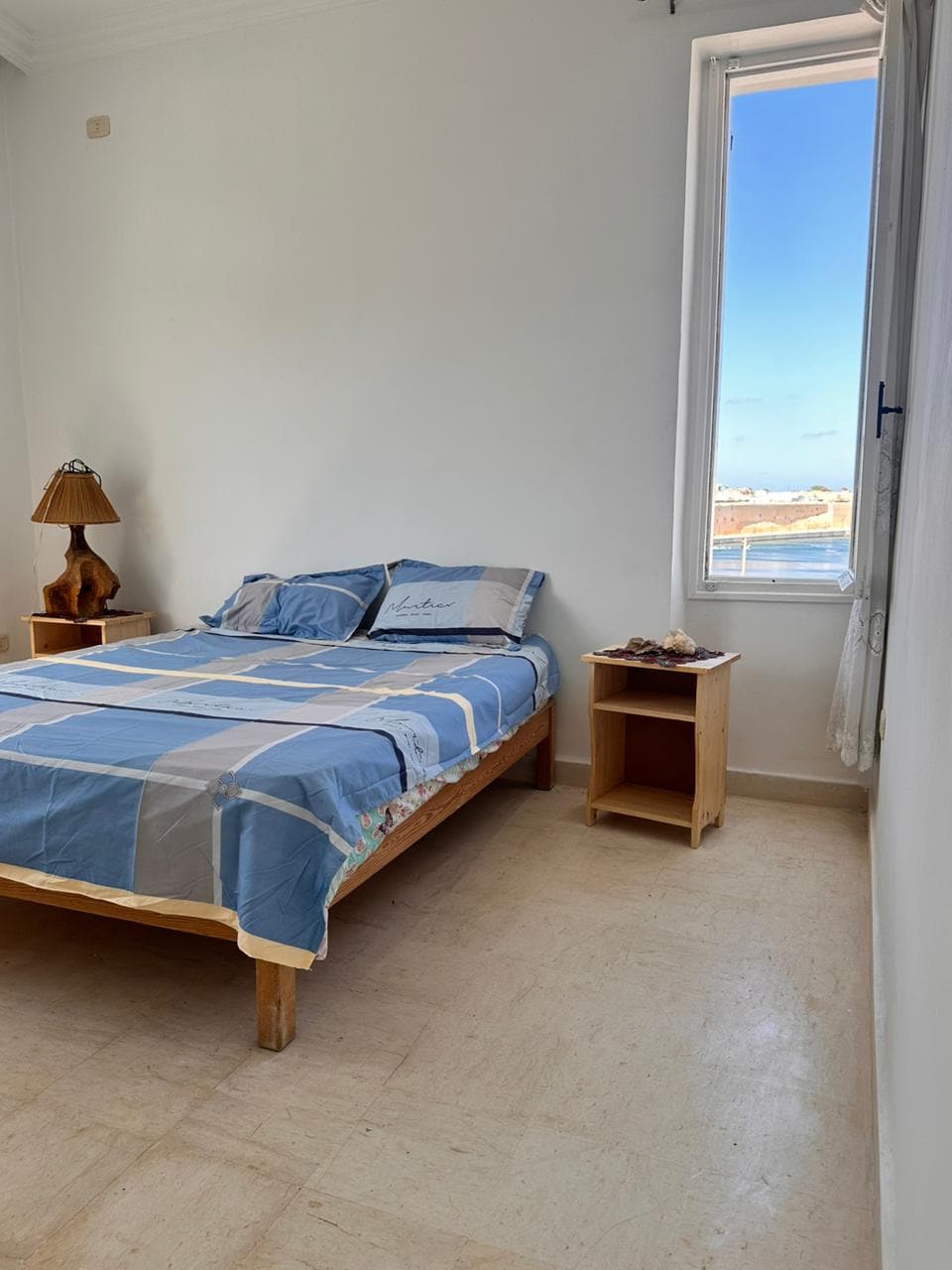 A spacious bedroom contains a bed dressed in a blue and white checked blanket. A bedside table holds a decorative lamp. Natural light enters through a window, highlighting the light-colored walls and floor.