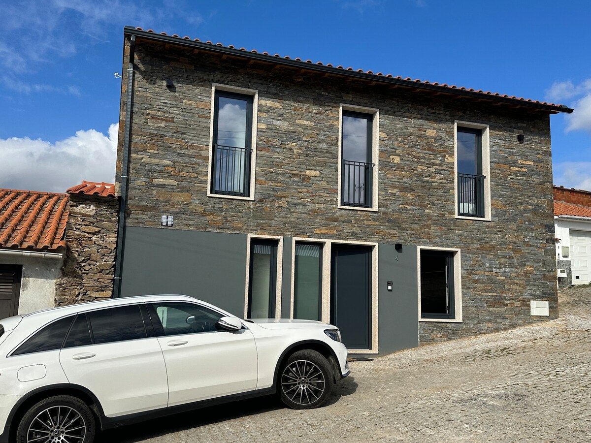 A rustic two-story house made of natural stone is shown. The façade features large windows with black railings and a modern grey entrance. A white vehicle is parked at the front, and the surrounding cobblestone pathway is visible under a partly cloudy sky.
