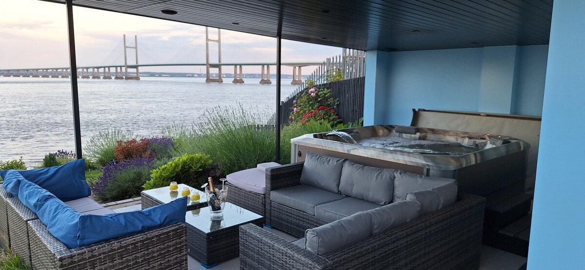 A shaded outdoor seating area features comfortable, cushioned seating surrounding a glass-topped table. A hot tub is positioned nearby, with views of the Severn Estuary and the bridge visible in the background. Lush greenery adds natural elements to the space.