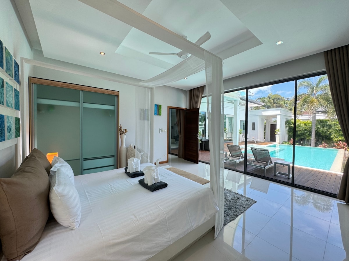 A serene bedroom features a spacious bed with neutral-toned linens, flanked by decorative pillows. Large glass doors provide a view of the private pool, while soft natural light brightens the white walls and polished tile floor.