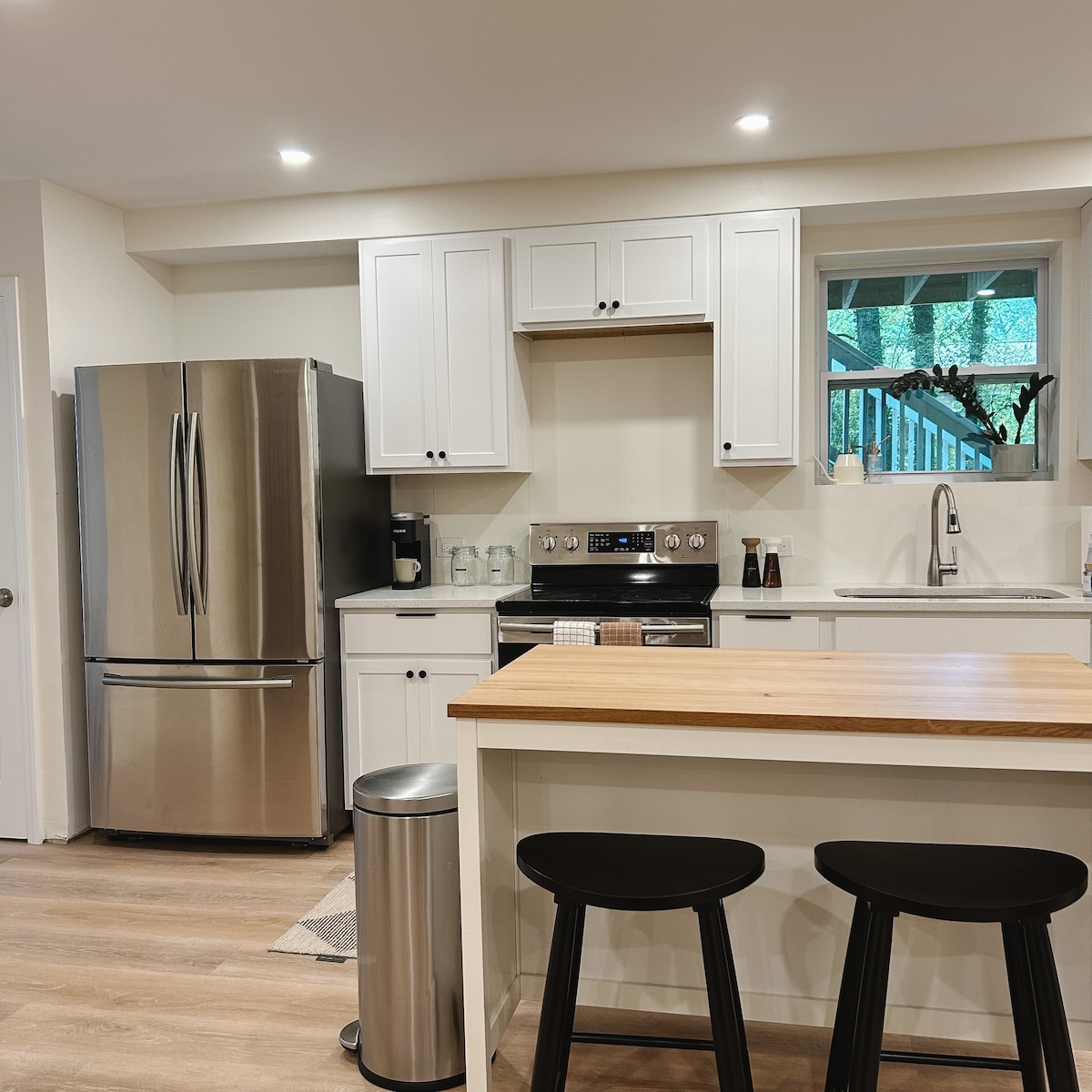 The kitchen features modern stainless steel appliances, including a spacious refrigerator and oven. A light wood countertop provides a practical prep space, complemented by two black bar stools positioned nearby. Natural light filters in through a window, enhancing the inviting atmosphere.
