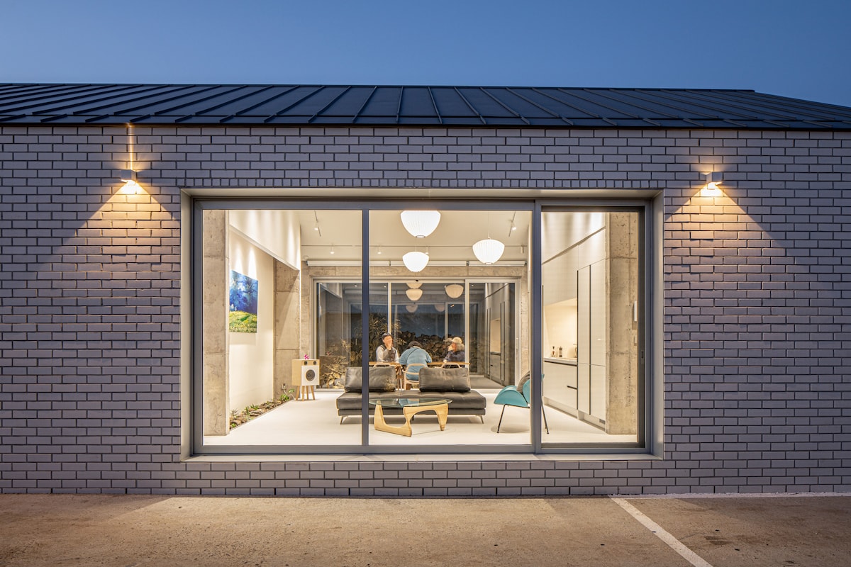 A modern exterior view of the accommodation showcases large glass windows reflecting soft outdoor lighting. Inside, a spacious living area is visible with a comfortable sofa and decorative elements that enhance the open layout, inviting guests to gather and relax.