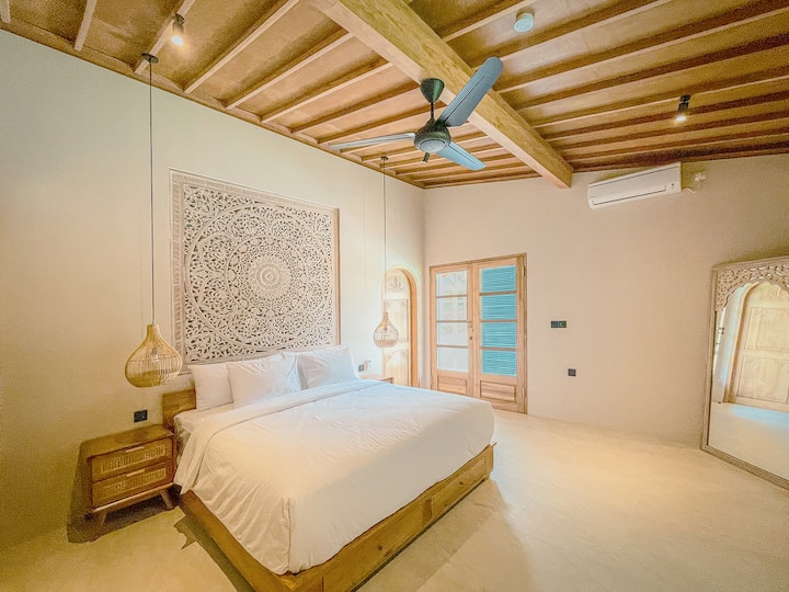 A spacious master bedroom featuring an ensuite bathroom & a king-size bed dressed in top-quality linens. It showcases a stunning hand-carved headboard, includes a wardrobe & is equipped with both air conditioning & a ceiling fan for ultimate comfort.