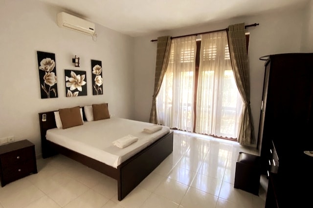 A spacious bedroom features a large bed with neutral bedding, complemented by decorative pillows. Elegant wall art depicting flowers is displayed above the bed. Natural light filters through the sheer curtains, and a dark wooden wardrobe and nightstands add to the room's functionality.
