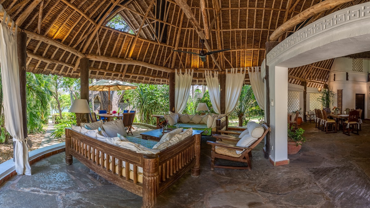 A spacious living area showcases natural wood beams and a thatched ceiling. Comfortable seating arrangements with light-colored cushions surround a low coffee table, while draped fabric provides a sense of privacy. Lush greenery is visible through large openings, enhancing the connection to the outdoors.